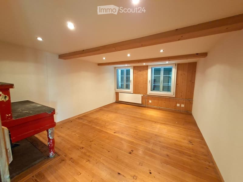 Empty room with wooden flooring, white walls, exposed wooden beams on the ceiling, and two windows with shutters.