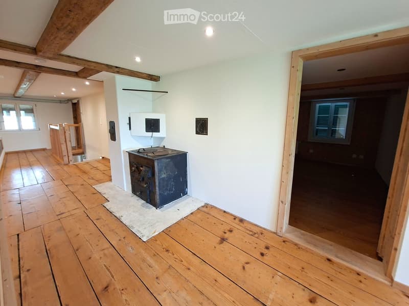 Empty room with wooden flooring, white walls, exposed wooden beams on the ceiling, and a small black stove in the center.