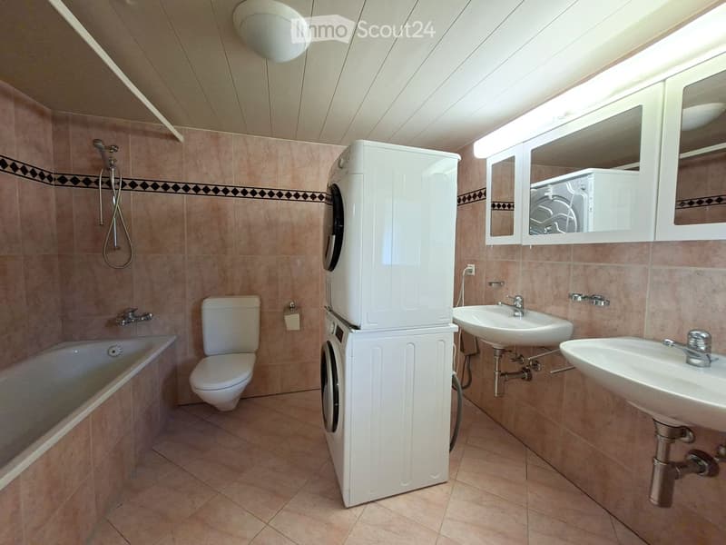 Bathroom with beige tiles, bathtub, toilet, two sinks, washing machine, and a mirror above the sinks.
