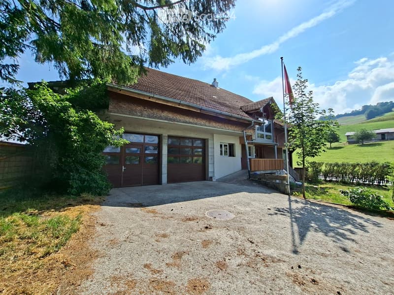 Two-story house with a red-tiled roof, large garage, and a driveway, surrounded by greenery and a flagpole.