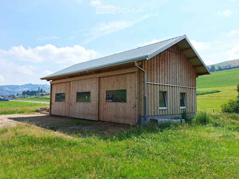 Single-story wooden house with a metal roof, large garage door, and multiple windows, situated in a green, rural landscape.