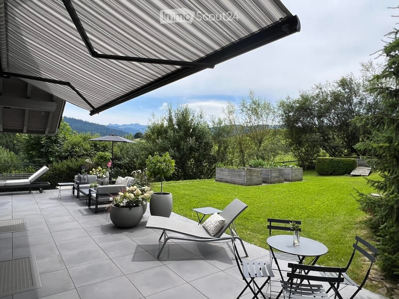 A modern outdoor patio with lounge chairs, small tables, and potted plants, overlooking a well-maintained lawn and scenic mountain backdrop.