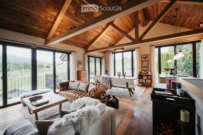 Modern living room with exposed wooden beams, large windows offering a scenic view, a brown leather sofa, a coffee table, an ottoman, a bench, a bookshelf, and a kitchen island.
