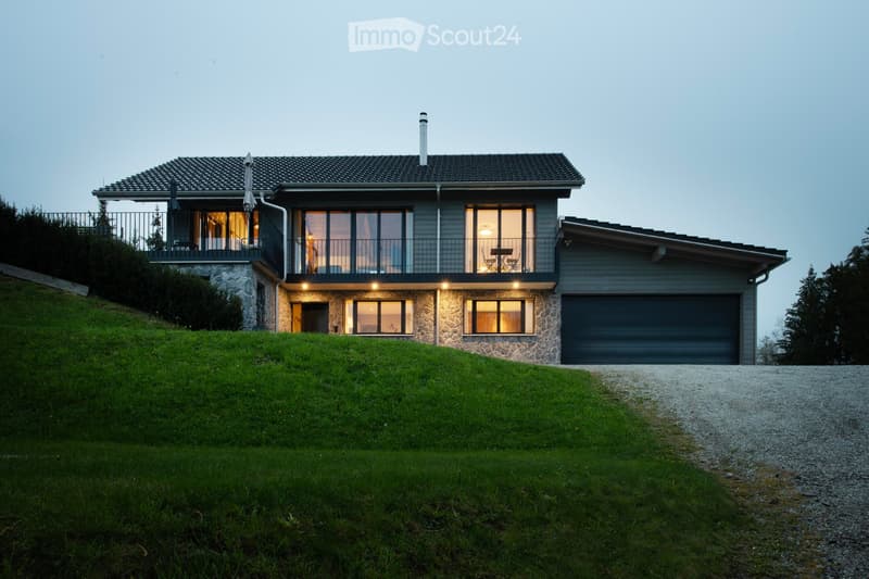 Modern two-story house with grey siding, stone accents, large windows, and a balcony on the upper level. Features a garage and is situated on a grassy slope with a gravel driveway.