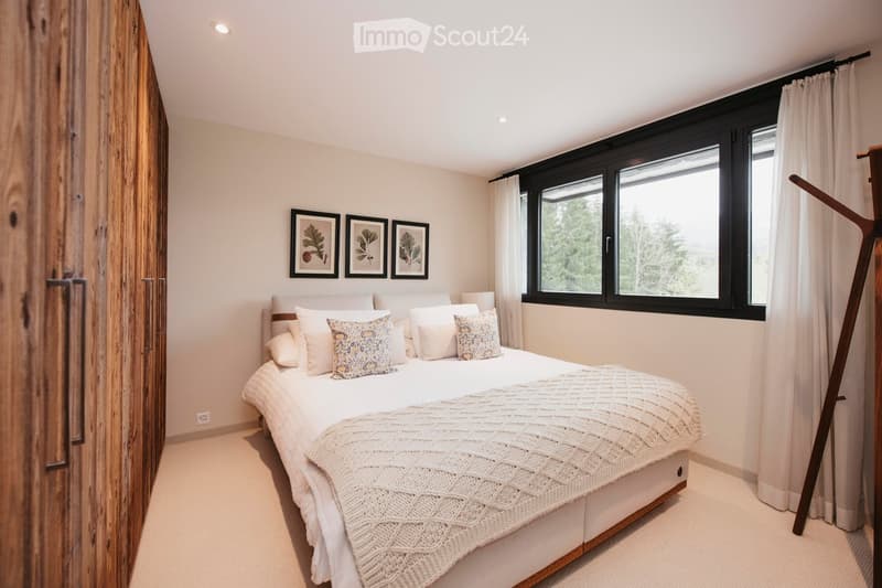 Modern bedroom with a large bed, decorative pillows, a knitted blanket, a wooden wardrobe, and three framed pictures above the bed. Large windows with curtains offer a view of greenery outside.