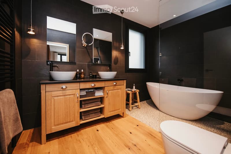 Modern bathroom with dark tiled walls, wooden floor, double sink vanity with wooden cabinet, bathtub, toilet, and a small wooden stool.