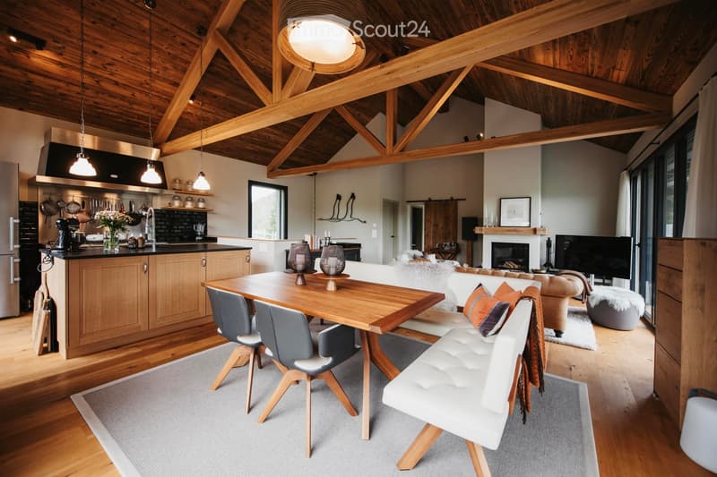 Open-concept living and dining area with exposed wooden beams, a modern kitchen, a dining table with chairs, a sofa, and a fireplace.
