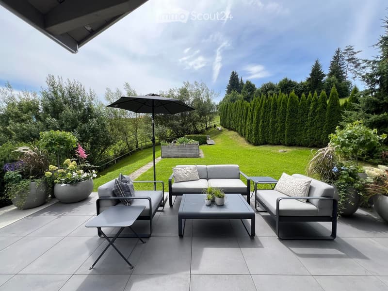 Modern outdoor patio with grey furniture, including a sofa, chairs, coffee table, and umbrella, surrounded by greenery and potted plants.