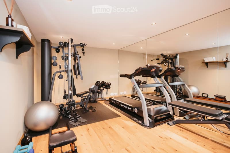 Well-equipped home gym with wooden flooring, large mirror, treadmill, elliptical machine, weight machine, exercise ball, and a small chair.
