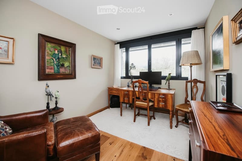 A well-furnished room with a leather sofa, wooden desk, chairs, a lamp, and decorative bird figurines. Large windows with white curtains allow natural light to fill the space.