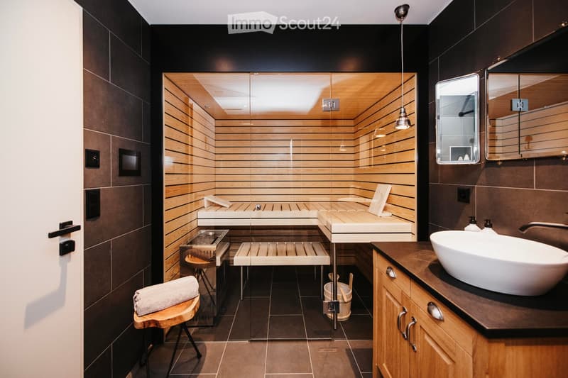 Modern bathroom with a sauna area, wooden benches, a sink, and a mirror. The sauna features wooden walls and a glass partition, with a towel on a wooden stool near the entrance.