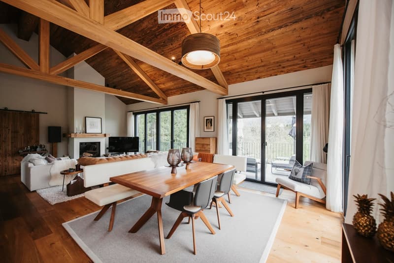 Modern living room with exposed wooden beams, large windows, a fireplace, a sofa, a dining table, and chairs.
