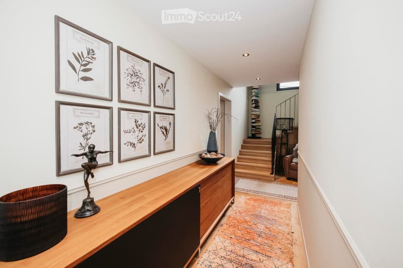 Elegant hallway with a wooden console table, framed botanical prints, a small sculpture, and a bowl of fruit. A staircase leads to an upper level, with a patterned rug and a seating area visible in the background.