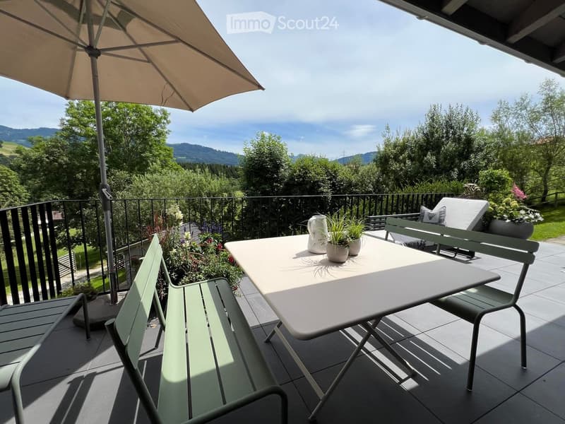 Balcony with a table, chairs, umbrella, and potted plants, overlooking a scenic mountain view.