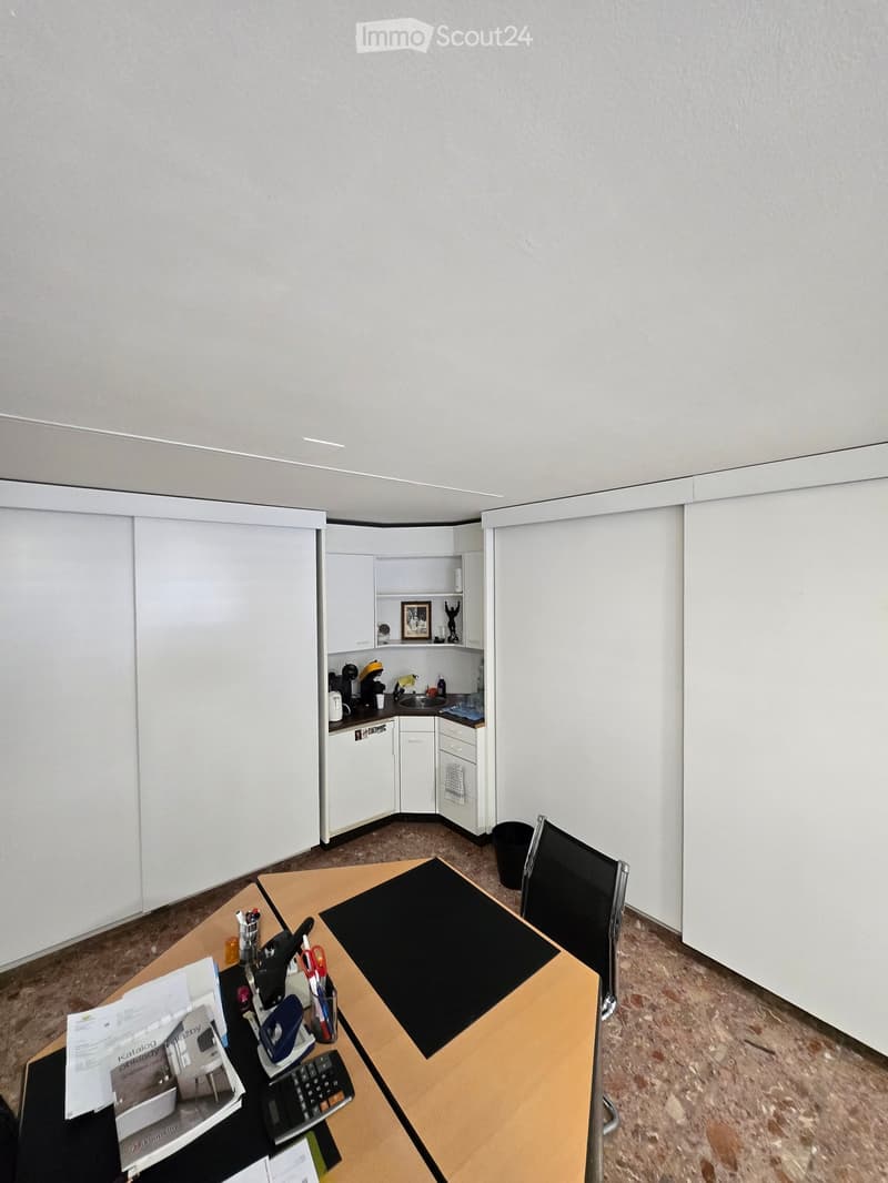 A modern, minimalist office space with a desk, chair, and a small kitchenette area featuring a fridge, sink, and cupboards.