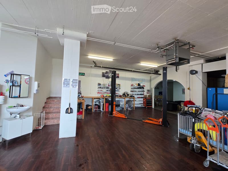 Large, open workshop or garage space with wooden flooring, white walls, and various tools and equipment. Includes a sink, tables, shelves, and a cart.