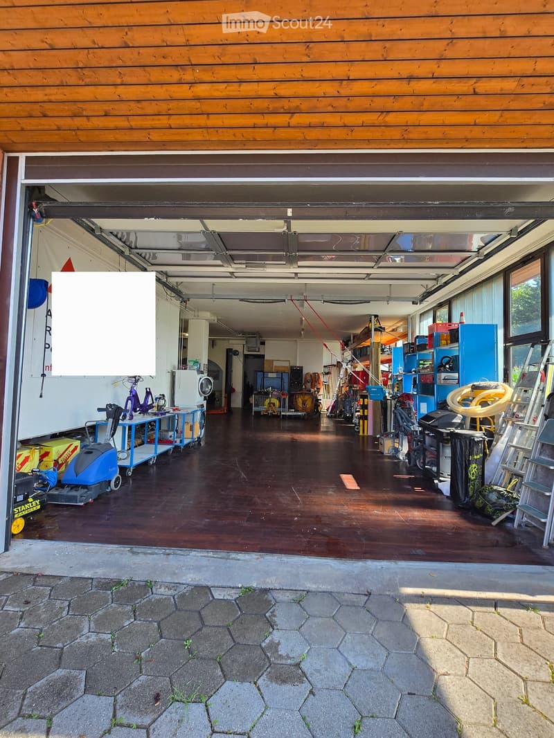 Open garage with wooden floor, various tools and equipment, large windows, and a partially open garage door leading to a paved outdoor area.