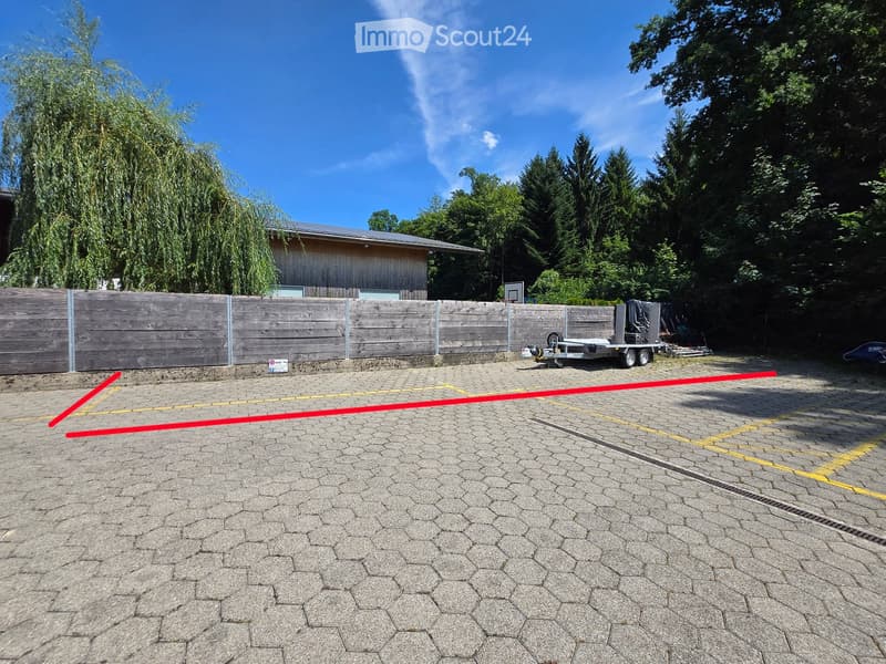 Paved outdoor area with marked parking spaces, wooden fence, trailer, and trees in the background.