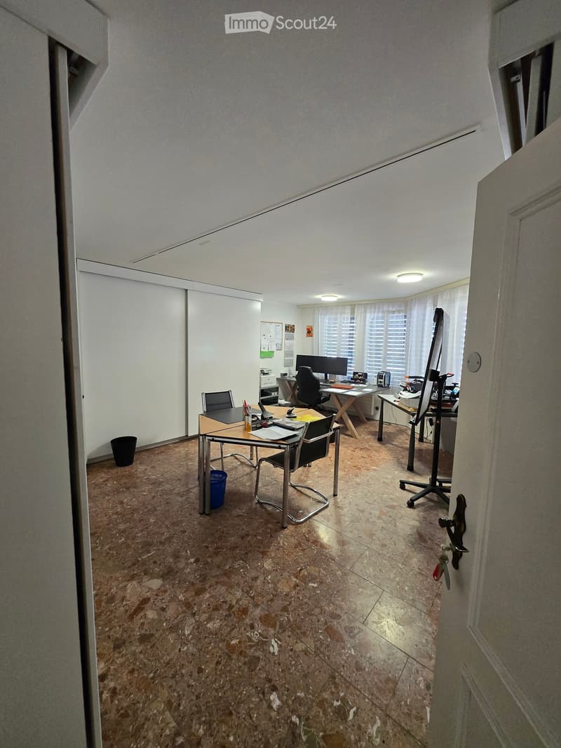 Office space with desks, chairs, computers, and office supplies. The room has a tiled floor and white walls.