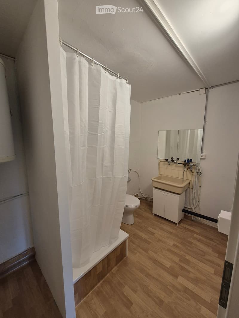 Bathroom with wooden flooring, bathtub with white curtain, toilet, sink with mirror, and small cabinet.