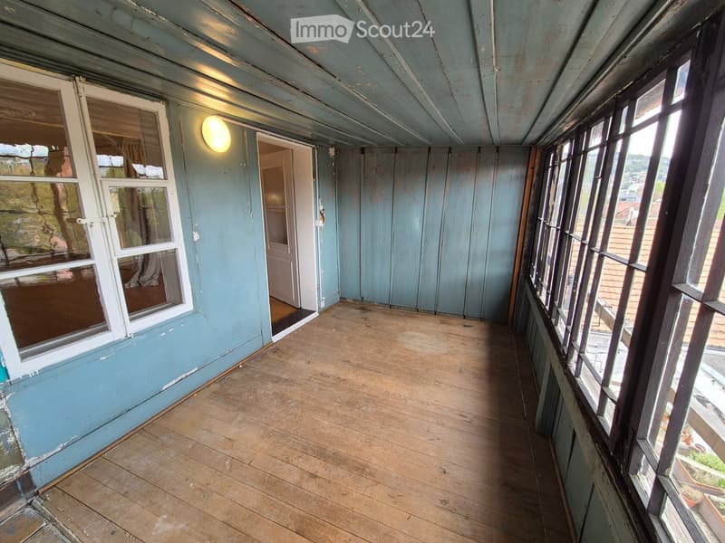 Empty wooden balcony with light blue walls, large windows, and a door leading inside.