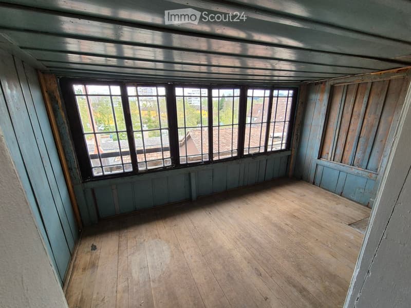Empty attic room with wooden floor, exposed wooden beams, and large windows offering a view of the city and rooftops.