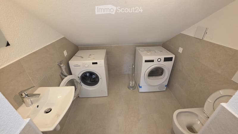 Modern bathroom with a sink, toilet, washing machine, and dryer. Beige tiled walls and floor, white fixtures.