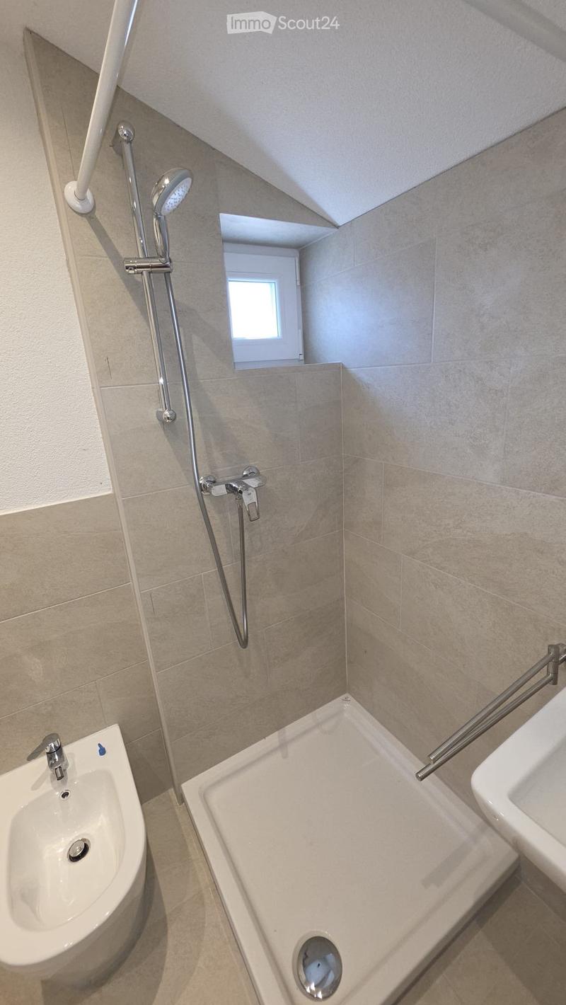 Modern bathroom with a bathtub, bidet, and showerhead, featuring beige tiles and a window for natural light.