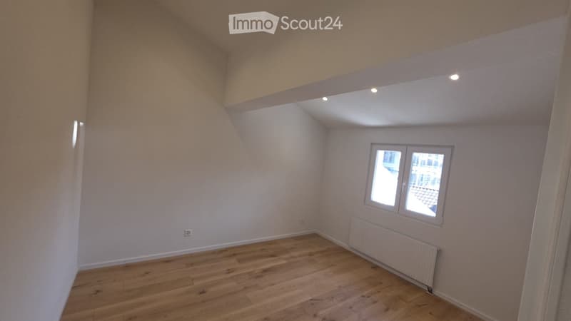 Empty room with wooden flooring, white walls, and a window with a view of the outside. The room features a slanted ceiling and built-in lighting.