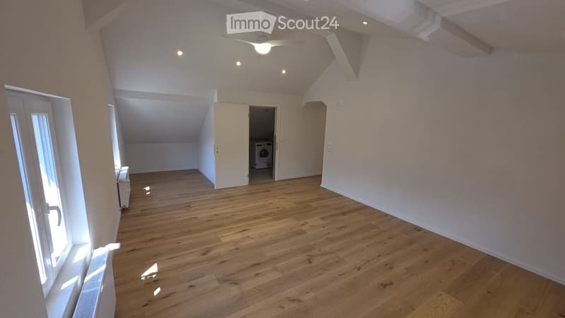 Empty room with wooden flooring, white walls, and a ceiling fan. A doorway leads to another room with a washing machine visible.
