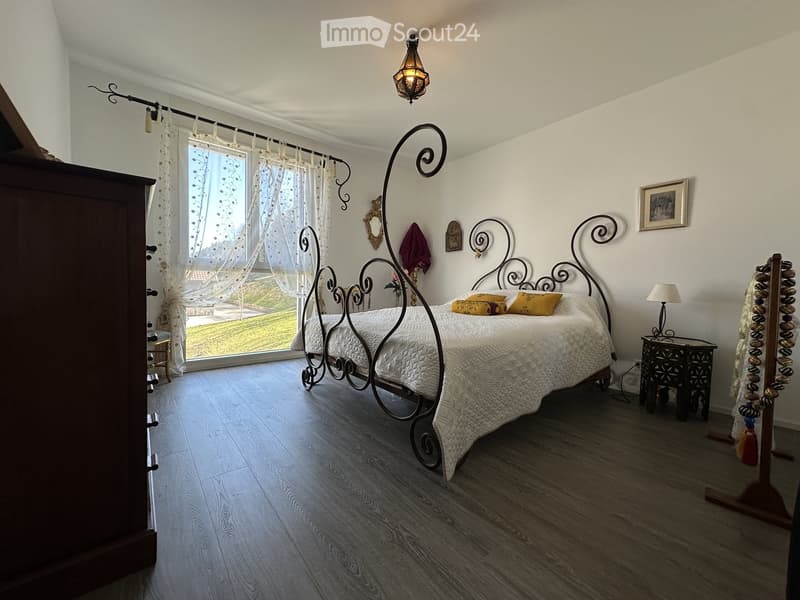 Elegant bedroom with a large, ornate metal bed, white bedding, wooden nightstand, and a view of a garden through large windows.