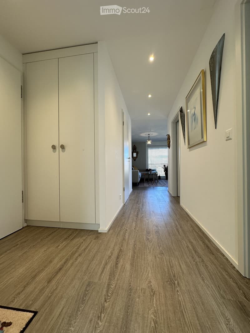 A well-lit hallway with wooden flooring, white walls, and several doors. Artwork is displayed on the walls, and a small rug is placed near the entrance.