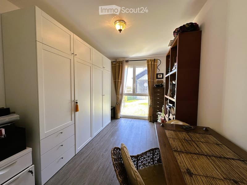Well-lit room with wooden flooring, large window with curtains, white built-in cabinetry, wooden table with chair, bookshelf, and decorative items.
