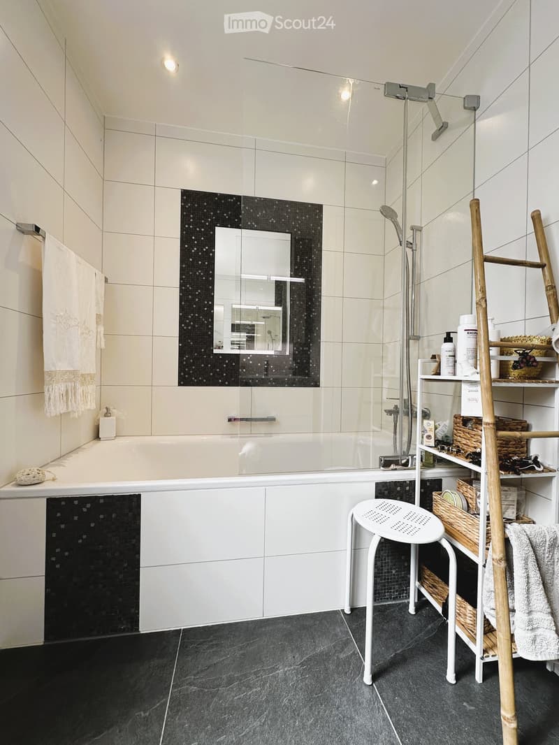 Modern bathroom with a bathtub, glass-enclosed shower, white tiles, black mosaic accents, a white stool, and a shelf with various toiletries and towels.