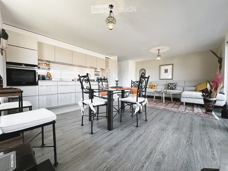 Open-plan living and dining area with a grey sofa, coffee table, dining table with chairs, and a modern kitchen with white cabinets and stainless steel appliances.