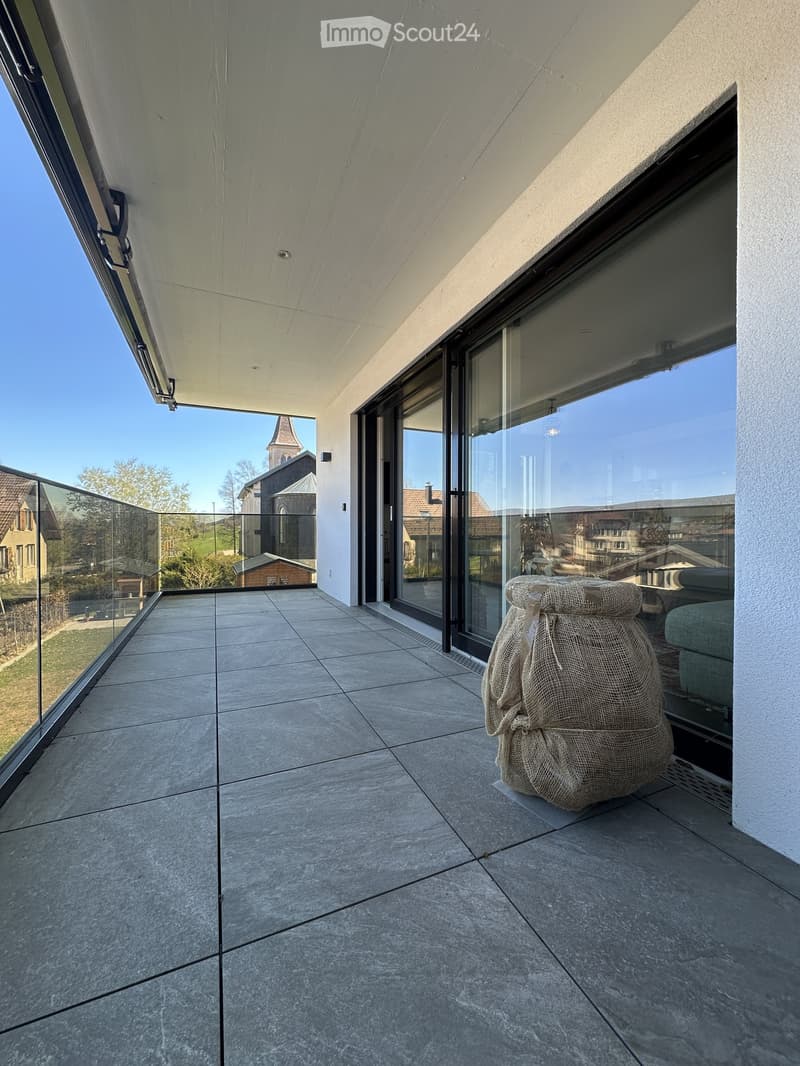 Modern balcony with large glass railings, tiled floor, and a view of the surrounding area including a church and houses.