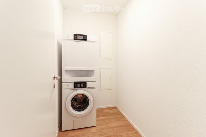 A small, empty room with white walls and wooden flooring, featuring a stacked washer and dryer unit against one wall.
