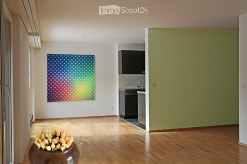 Bright, open living space with wooden flooring, a colorful abstract painting on the wall, a vase with flowers, and a compact kitchen area with cabinets and appliances.