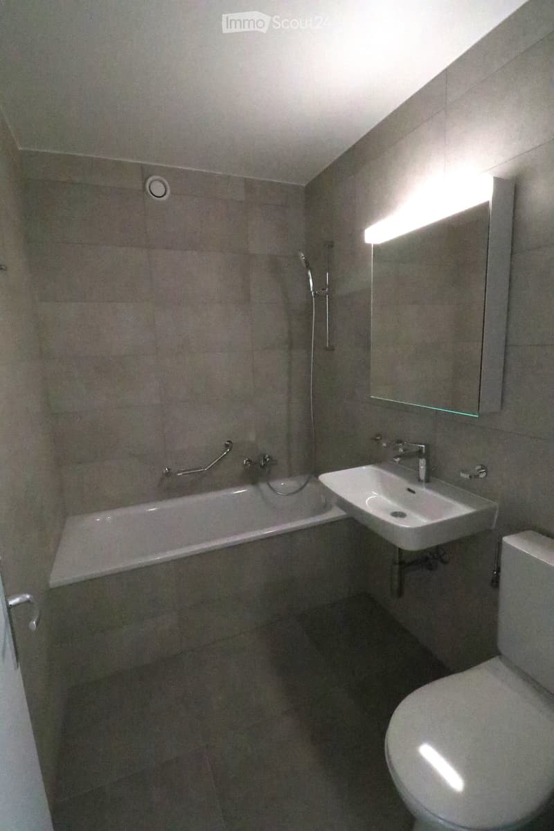 Modern bathroom with a bathtub, sink, toilet, and mirror. The walls and floor are tiled in a neutral color scheme, and the space is well-lit with natural light coming from a window.