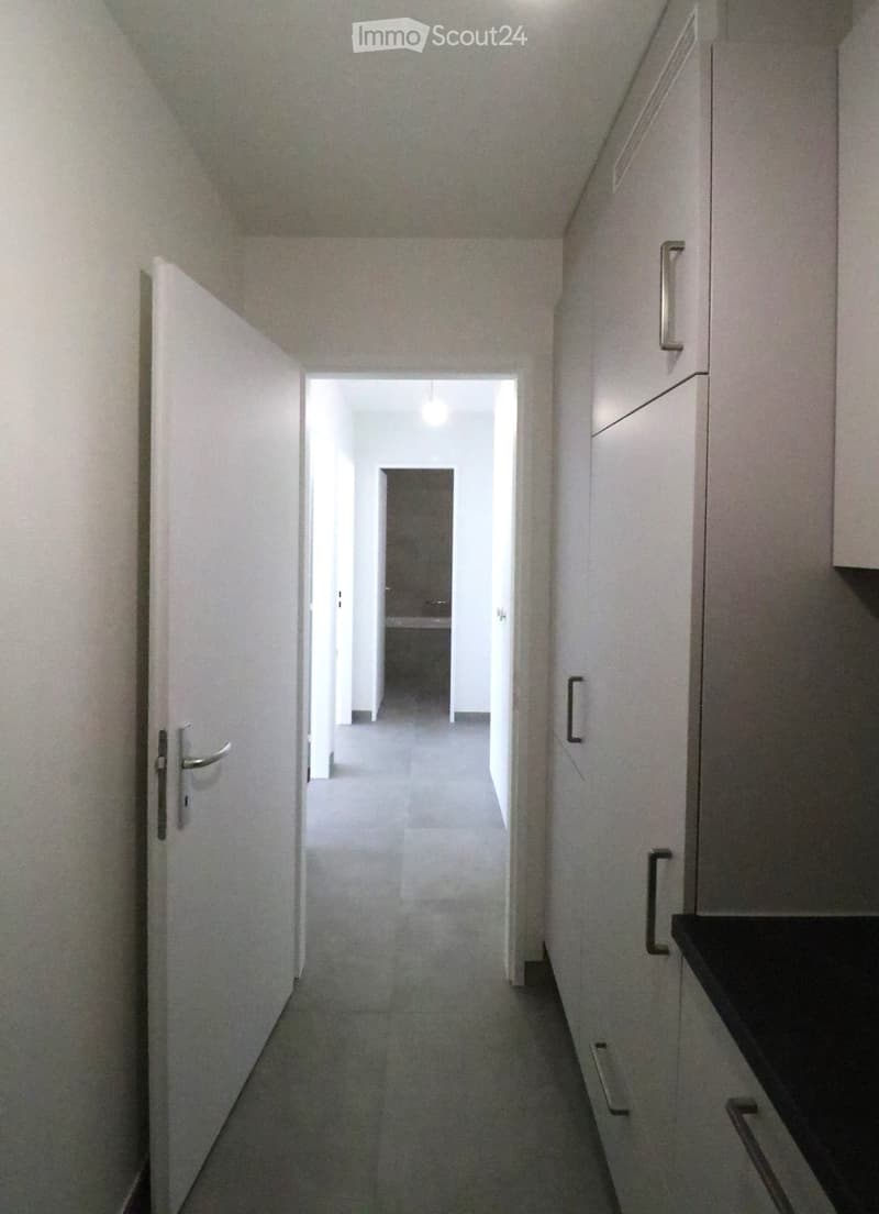 A narrow, well-lit hallway with white walls, grey tiled floor, and white cabinetry on one side. The hallway leads to an open doorway at the end.
