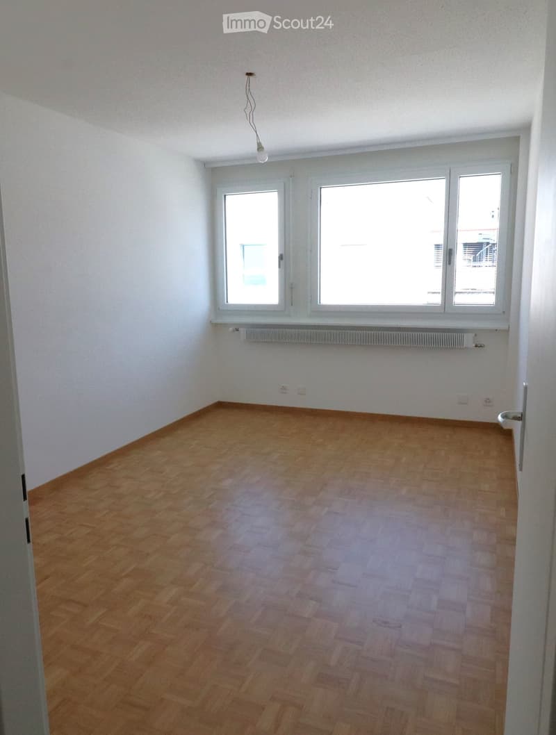 Empty room with wooden flooring, white walls, and large windows allowing natural light. The room features a radiator below the windows and a single light fixture on the ceiling.