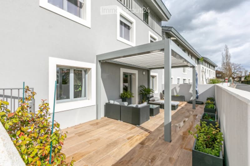 Modern outdoor patio with wooden decking, grey furniture, pergola, and potted plants.