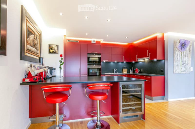 Modern kitchen with red cabinetry, built-in oven, wine cooler, island with bar stools, coffee maker, and framed artwork on the walls.