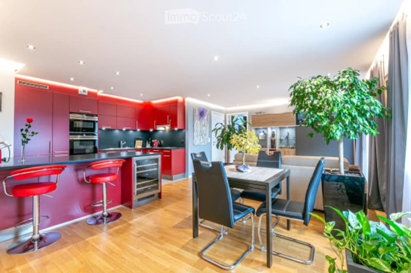 Modern open-plan kitchen and dining area with red cabinets, stainless steel appliances, red bar stools, a dining table with chairs, and potted plants.