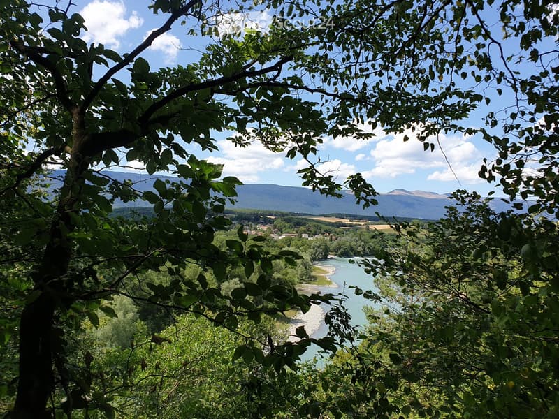 A scenic outdoor view showing a river winding through a valley, surrounded by lush green trees and distant mountains under a partly cloudy sky.