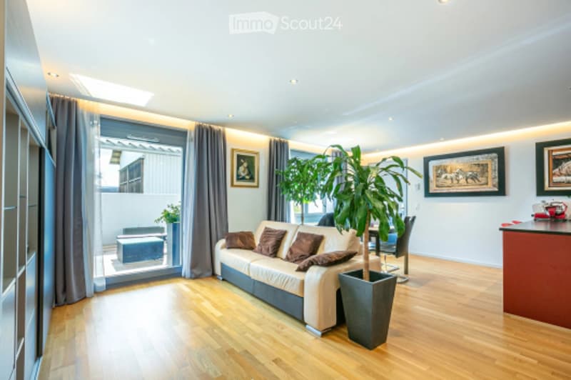 Bright living room with wooden flooring, large windows, a cream sofa with brown throw pillows, a coffee table, a potted plant, and a bar counter.