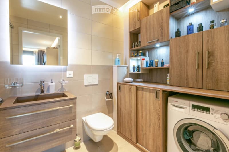 Modern bathroom with wooden cabinets, a sink, toilet, and a washing machine. The space features a large mirror, a faucet, and various toiletries on the shelves.