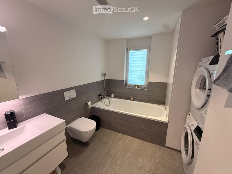 Modern bathroom with a white sink, toilet, bathtub, and a washing machine. Features a window with blinds and a grey accent wall.