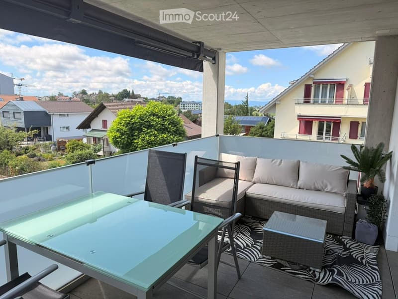 Balcony with a modern glass table, wicker sofa, coffee table, and potted plant, overlooking a residential area with houses and greenery.