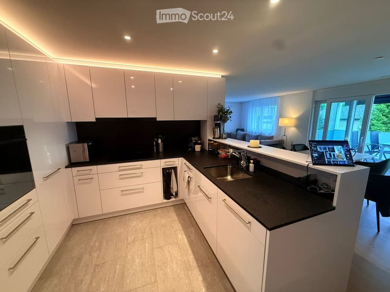 Modern kitchen with white cabinets, black countertops, and integrated appliances. Features a central island with a sink and a laptop on the counter.